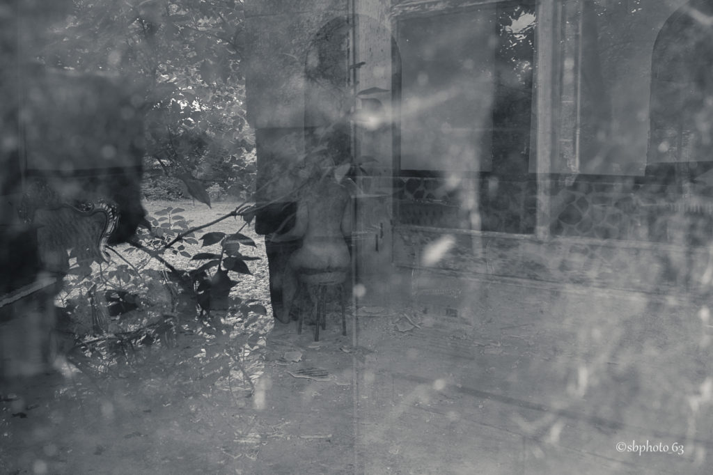 photo en noir et blanc prise au travers d'une fenetre, on y voit une femme nue assise de dos