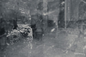 photo en noir et blanc prise au travers d'une fenetre, on y voit une femme nue assise de dos