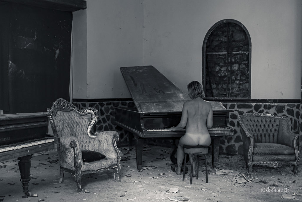 photo en noir et blanc d'une femme de dos jouant au piano nue dans un endroit à l'abandon