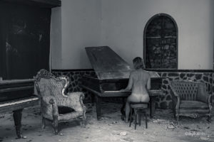photo en noir et blanc d'une femme de dos jouant au piano nue dans un endroit à l'abandon