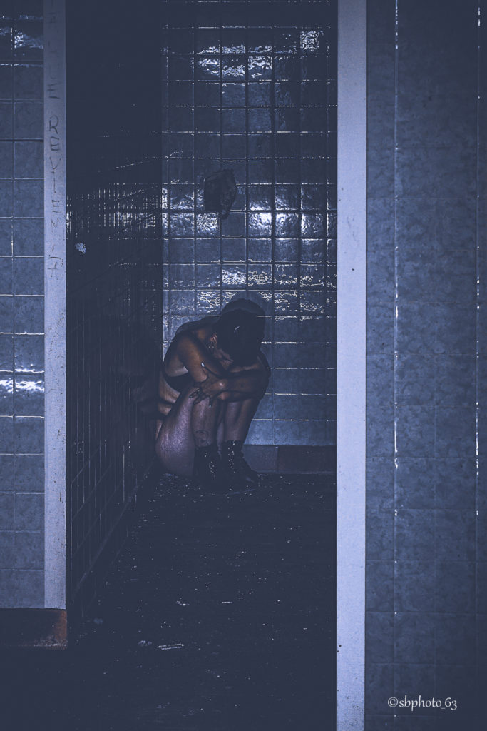 photo en couleur, avec une tendance bleutée, d'une femme recroquevillée dans une ancienne douche d'un ancien hopital