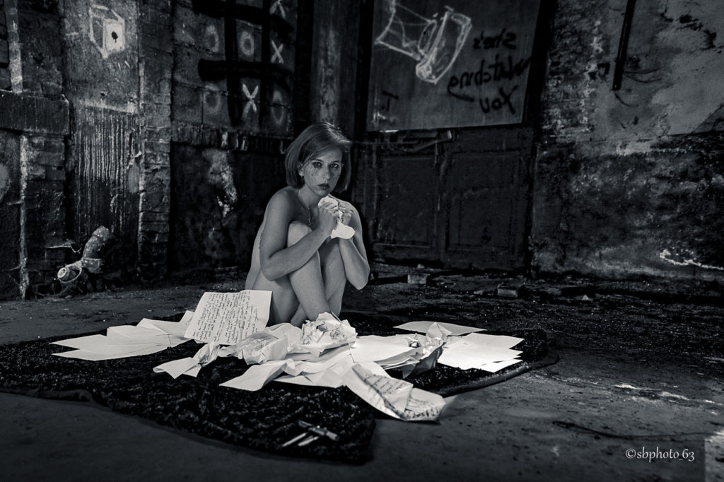 photo en noir et blanc d'une femme dans un lieu urbex. elle est assise dans les feuilles, recroquevillée elle serre dans feuilles blanches dans ses mains