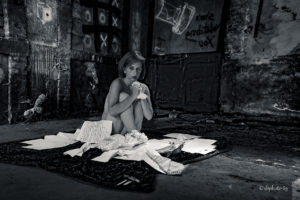 photo en noir et blanc d'une femme dans un lieu urbex. elle est assise dans les feuilles, recroquevillée elle serre dans feuilles blanches dans ses mains