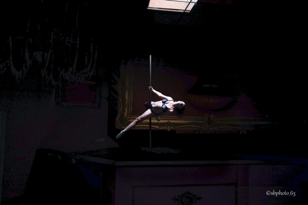 photo d'une femme en train de réaliser une figure de pole dance, elle est sous un puit de lumière. Cette photo couleur est sombre hormis le corps de cette femme et la barre de pole dance