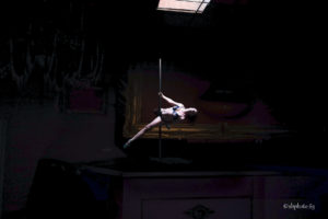 photo d'une femme en train de réaliser une figure de pole dance, elle est sous un puit de lumière. Cette photo couleur est sombre hormis le corps de cette femme et la barre de pole dance