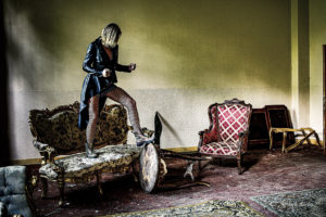 photo couelur d'une femme dans un lieu abandonné, elle se tiend debout sur un ancin sofa, elle est en cuir, bas résille et doc martens