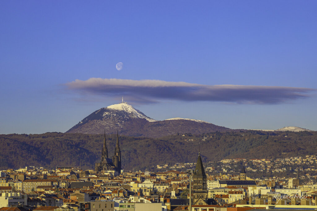 C'est une vue du paysage Clermontois, elle se décompose en trois plans. Le premier plan est consacré à l'urbanisme, notamment les toitures. Le deuxième plan la cathédrale de Clermont-Ferrand, et ses célèbres flèches. Et au troisièmement plan le Puy-de-Dôme. Sur monté d'un banc de nuages qui lui-même est surmonté de la lune, tout ce qui se passe dans un ciel bleu.